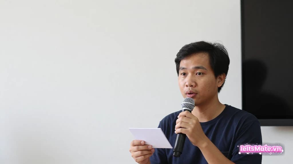 Một người học Việt Nam cầm thẻ câu hỏi trắng trước micro nhỏ, tạm dừng suy nghĩ trước khi trả lời.