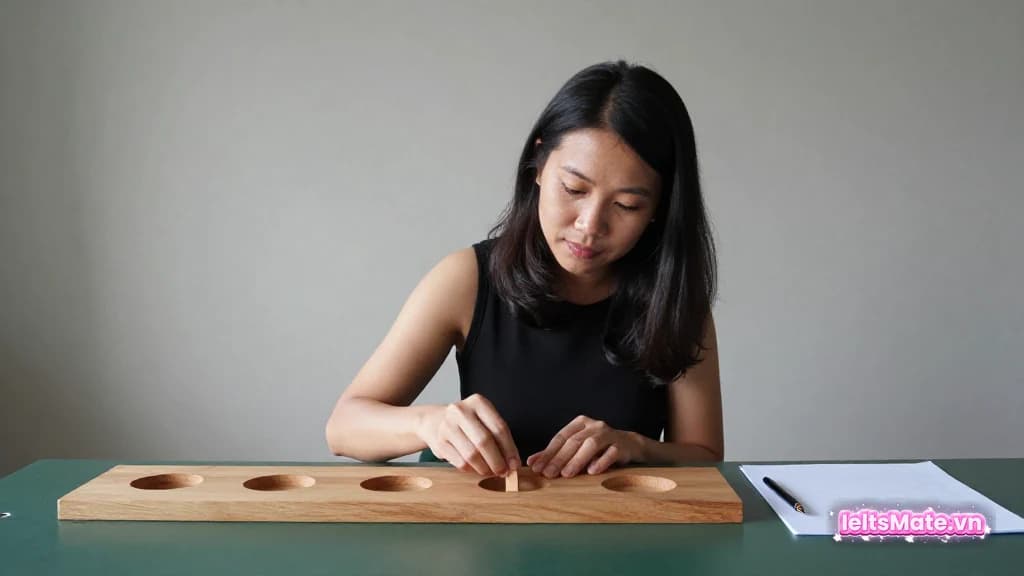 Một người học Việt Nam kiểm tra năm ô trống trên bảng gỗ cạnh trang giấy trắng và bút trên bàn.